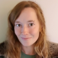 A woman with long wavy blonde hair, wearing a green top and fur-trimmed jacket, smiles at the camera. She has a small facial piercing above her right eyebrow. The background is plain and light-coloured.