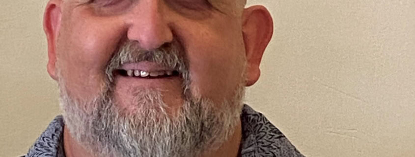 A middle-aged man with a bald head, gray beard, and mustache smiles at the camera. He is wearing a patterned gray shirt and is positioned in front of a plain, light-colored wall.