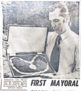 A man in a shirt and tie holds an open case displaying a ceremonial chain of office. The newspaper caption identifies him as the Carnival Committee secretary presenting the chain to the town’s first mayor.