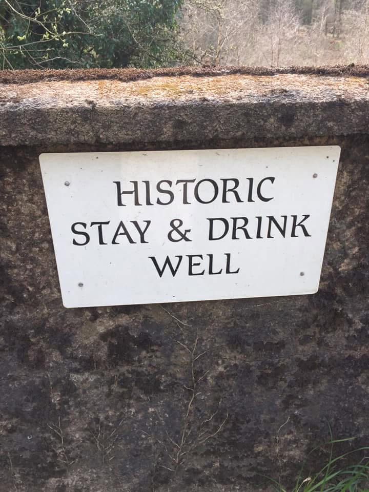 A white rectangular sign attached to a weathered stone wall that reads, “HISTORIC STAY & DRINK WELL.”